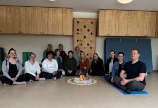 Die Eltern der Kindergartenkinder sitzen mit der Yogalehrerin im Halbkreis auf dem Boden der Turnhalle