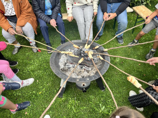 Menschen sitzen um eine Feuerschale und backen Stockbrot.