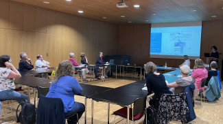 14 Menschen sitzen an Tischen und verfolgen eine PowerPoint-Präsentation