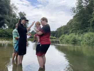 Pfarrerin Popp steht im Wasser der Rott und tauft ein Baby. Es wird vom Taufpaten gehalten. Im Hintergrund sieht man den Fluss mit ufergesäumten Bäumen.